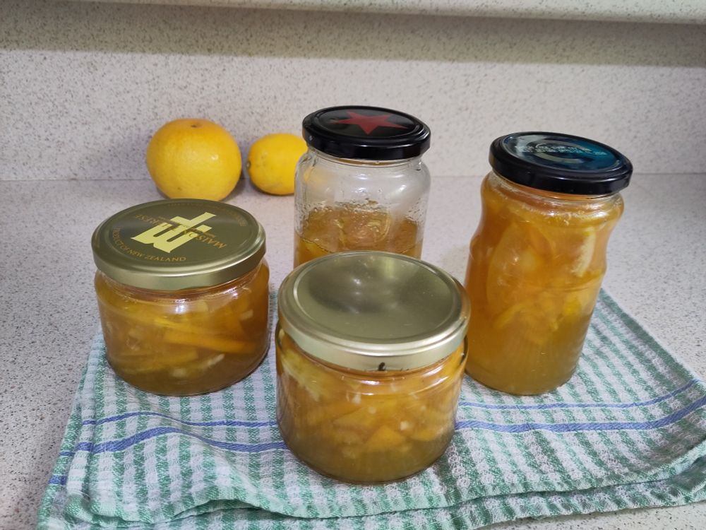 4 glass jars of marmalade - a dark orange with the paler bits of skin towards the top of each jar. The front two squatter jars have copperish, the two taller, thing jars towads the back have black lids. They are sitting on a folded green and white checker tea towel. In the background are a grapefruit and lemon (both yellow). Everything is resting on a specked bench top.