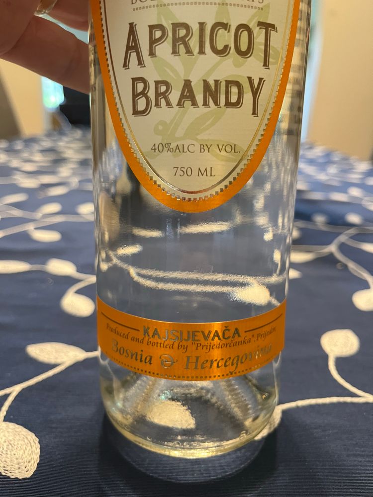 Bottle of clear apricot brandy with an orange label that’s a little hard to see but says “kajsijevača, produced and bottled by Prijedorčanka, Prijedor, Bosnia and Hercegovina