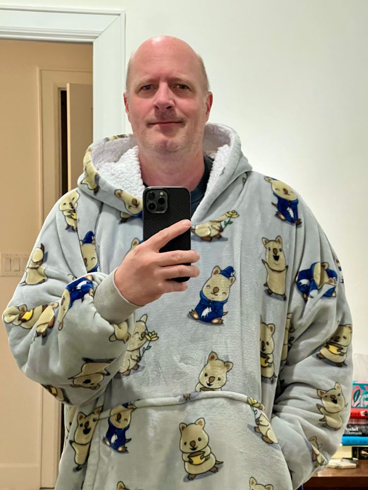 Selfie of a bald middle aged guy standing in a white room wearing an enormous oversized fuzzy gray fleece-lined hoodie covered in happy cartoon quoakkas