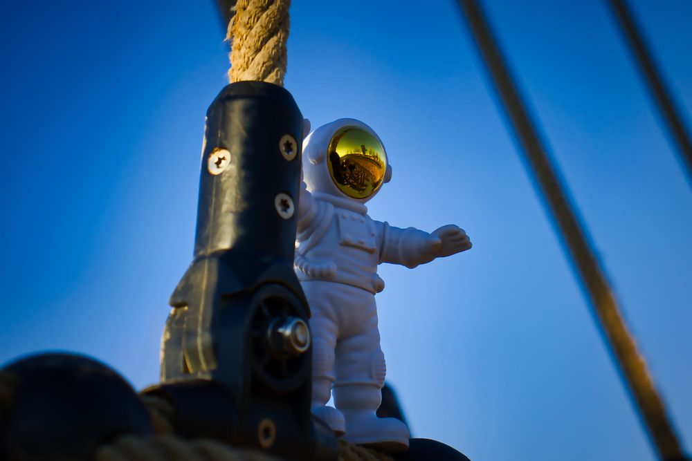Ein weißer Astronaut mit goldenem Visier (Figur) steht auf einem Gebilde aus verschiedenen Werkstoffen, Gelenken, Tau, Schrauben. Im Hintergrund blauer wolkenloser Himmel