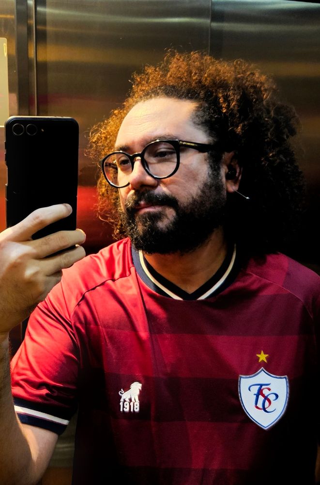 Selfie no espelho de um elevador. Homem de barba, óculos redondo, usa uma blusa vermelha do Fortaleza com símbolo retrô. Cabelo grande e cacheado preso pra trás em uma tiara. 
