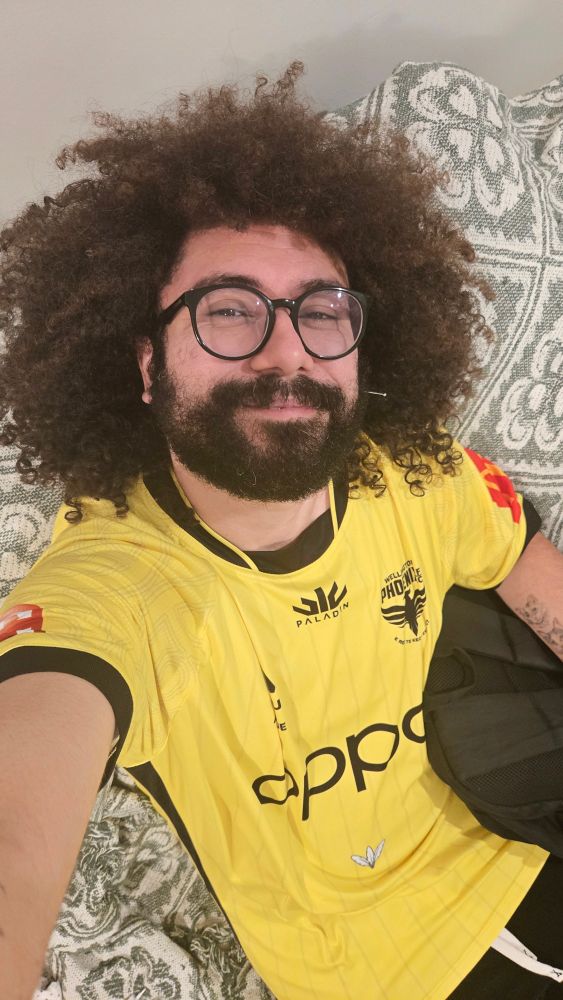 Selfie. Homem de cabelo grande cacheado, usa óculos redondos e tem barba. Tá sentando em um sofá. Ele usa uma blusa amarela do time Wellington, da Nova Zelândia.