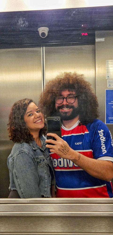 Casal tirando foto no espelho. Ela usa jaqueta jeans azul, ele uma blusa azul, vermelha e branca do time do Fortaleza.