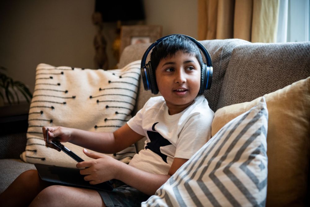 A young boy sitting on a sofa wearing headphones and holding a tablet. He is looking round smiling at someone not seen in the photo.