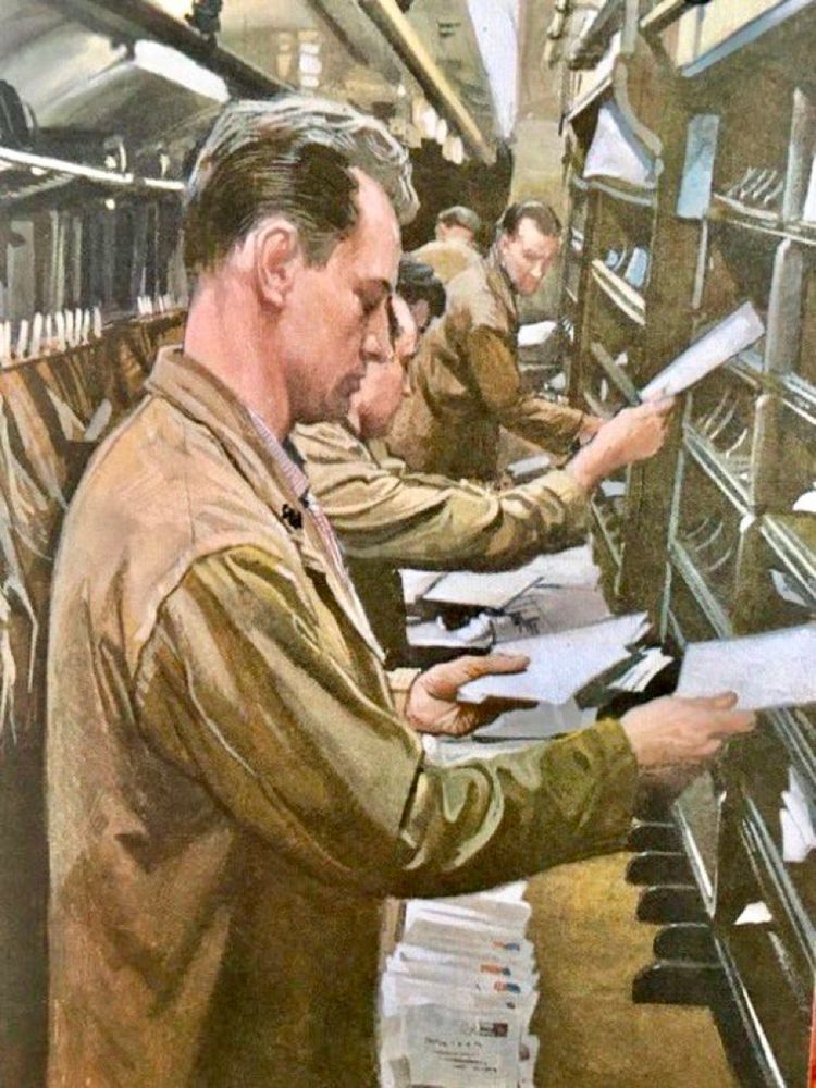 A column of men with receding headlines and sharp partings stand sorting mail into pigeon holes.
They are standing in a long thin chamber: a railway carriage adapted for mail. The men are all wearing sandy coloured coats to protect their clothes.