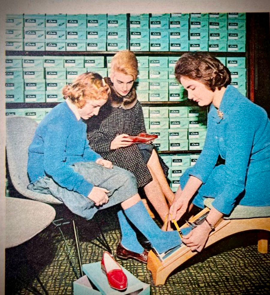 A 1960s photograph of a smartly dressed mother and child in a shoe shop.
The child puts her socked foot on the measuring slide on the sloping footstool while an assistant, flanked by boxes and boxes of Clarks shoes, fits the measuring strap