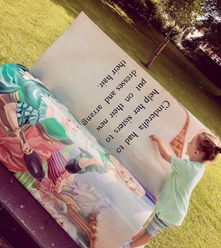 Photograph taken at an angle, showing a young child, perhaps four years old, pointing to a park bench created to look like an open ladybird book (Cinderella)