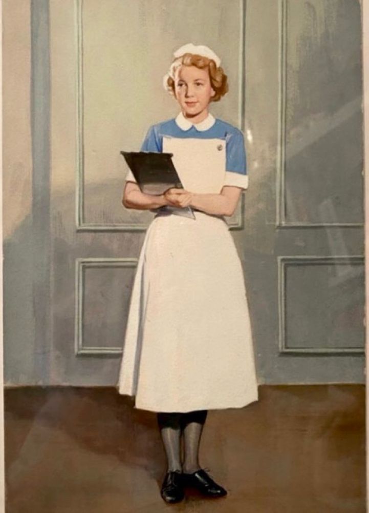 A full length portrait of a young, female nurse holding a clipboard. She is in her traditional outfit with crisp white pinafore, black tights and shiny flat black shoes. She is looking straight ahead, a little shyly. 