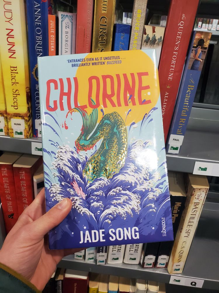 A picture of the novel Chlorine, by Jade Song which portrays a bloody fish tail in splashing water, against a backdrop of library books.
