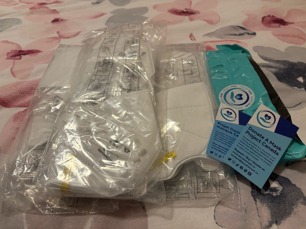 A pile of free sample packs of respirator style masks on a floral background. Beside the masks are business cards form Donate A Mask Project Canada. 