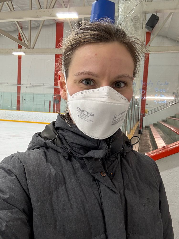 A white woman smiles to the camera in a white Vitacore N95 respirator. She is at a skating rink and the ice and part of the stands are visible behind her. She is also wearing a grey jacket, and her hair is out of place from her pony tail. 