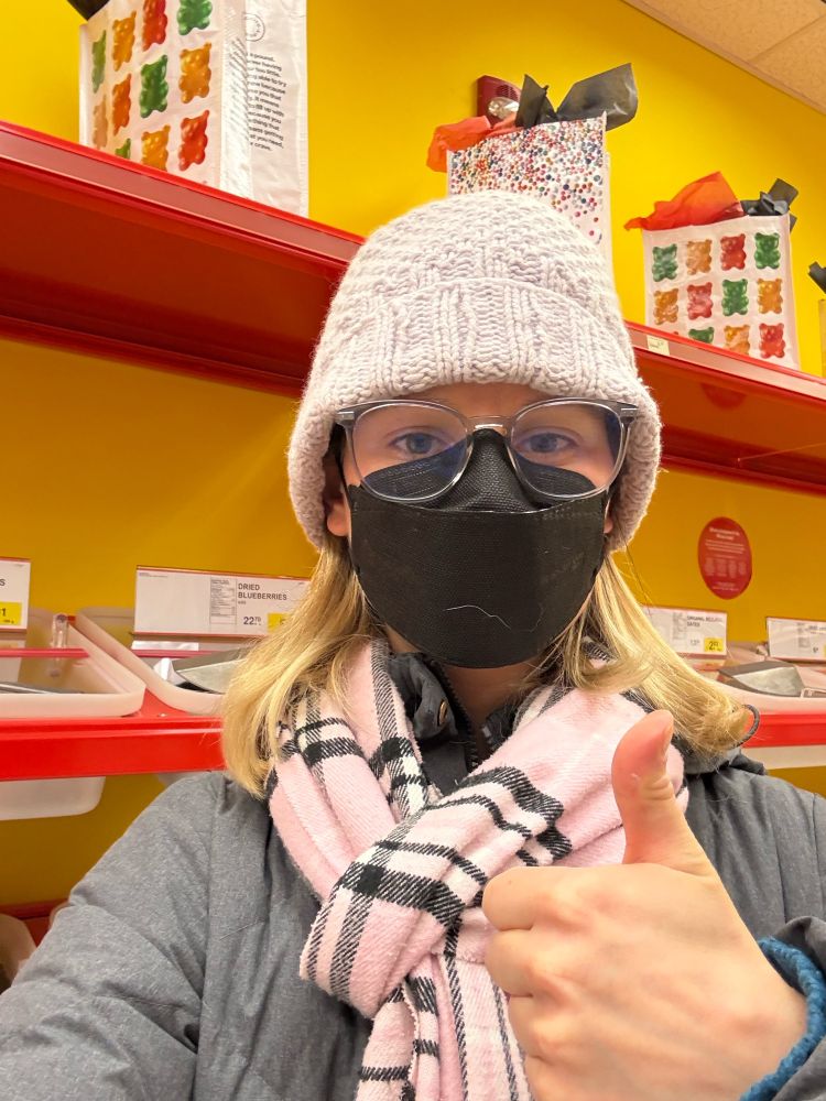 A white woman holds a thumbs up in a bulk food store. She is wearing a lilac knitted toque, glasses, a black KN95 mask, a pink scarf and a grey winter jacket. The wall behind is yellow with red shelving containing food in bulk bins. 