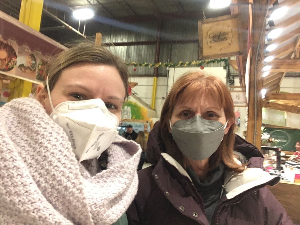 Selfie of two women, a mother and daughter, dressed in winter jackets and wearing N95 style respirators in a warehouse style farmers market. 