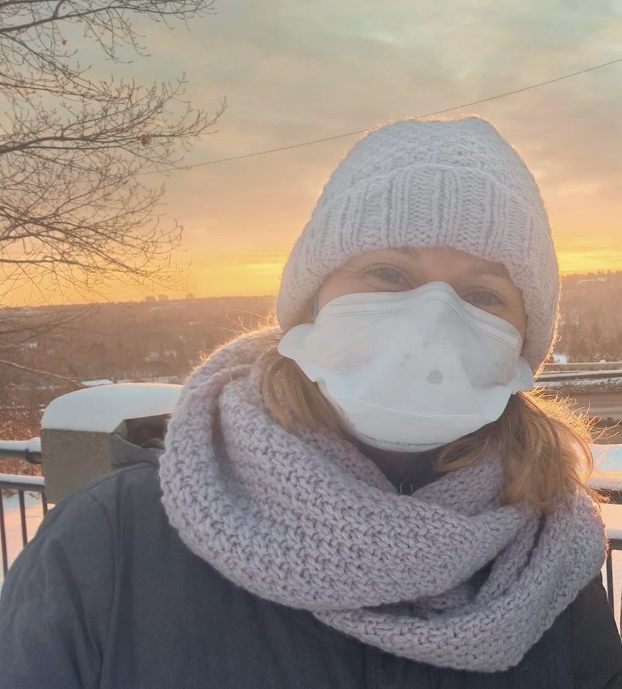 Selfie of a white woman against a winter sunset. She is wearing a lilac knitted toque and matching scarf, a white duck bill style respirator and a grey winter jacket. She is posing above a flat city scape with mostly trees and a cluster of three tall buildings far in the horizon. The sun is low, making the top of the sky a pinkish-orange, brightening toward yellow at the horizon. There is snow on the railing behind. 