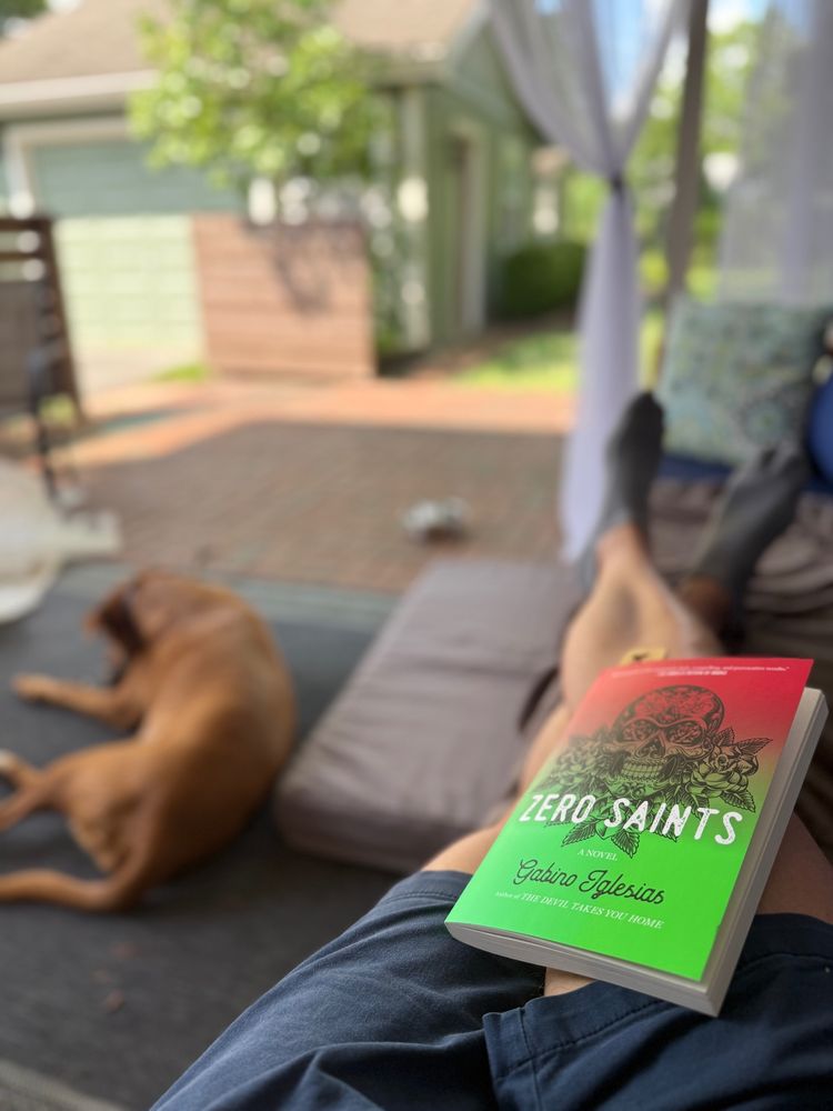 Photo of Zero Saints novel being read on a patio with a dog lying below.  