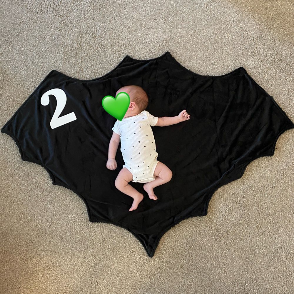 Baby girl in a white onesie with black triangles laying on a fuzzy black bat-shaped blanket with a white 3D printed number 2.