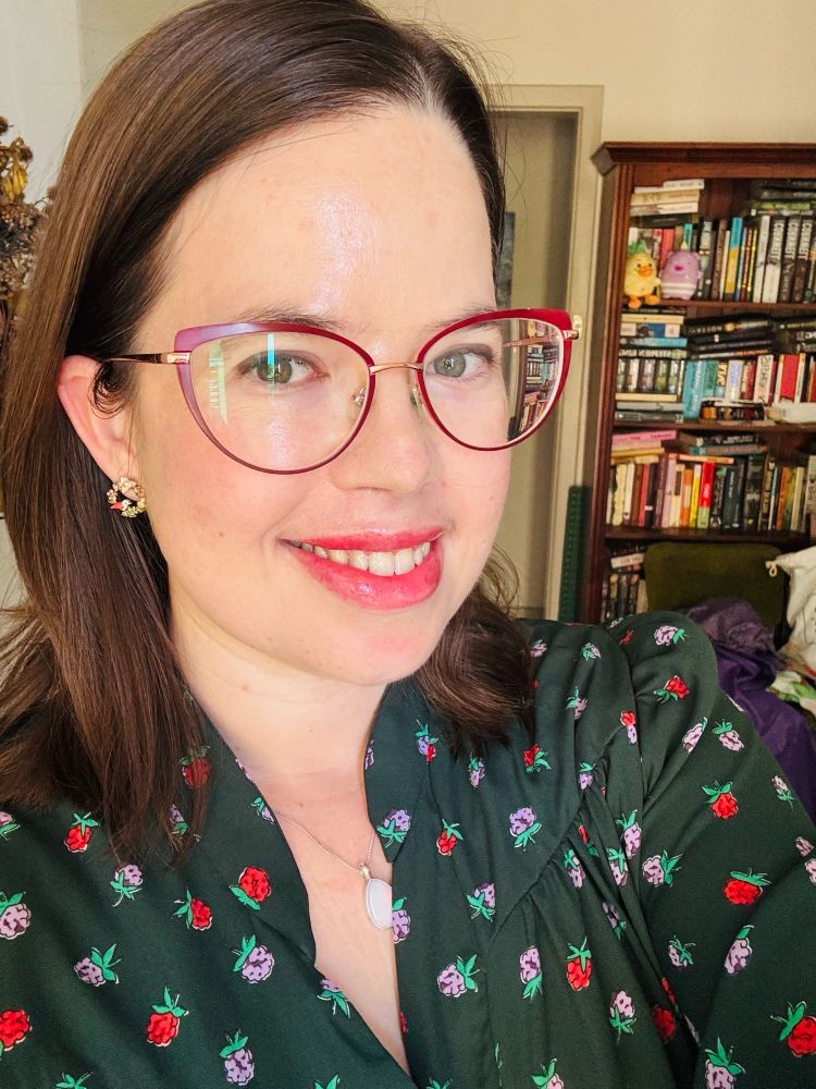 A woman wearing red rimmed glasses smiles at the camera. She’s wearing a dark green dress with a pattern of red flowers and purple flowers. She’s dressed up for Christmas lunch. 