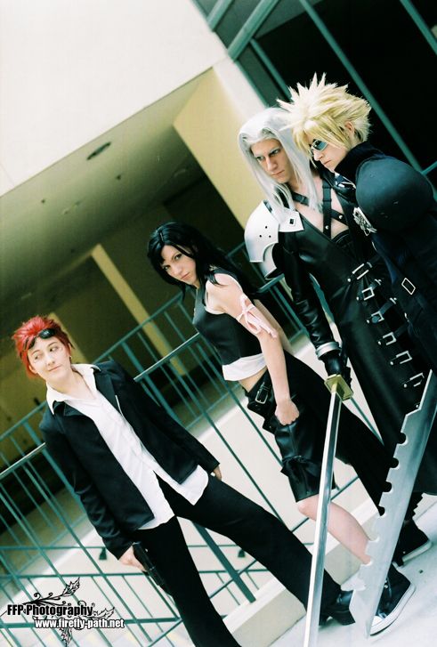 four cosplayers stand side by side dressed as final fantasy vii characters in various intricate outfits made of dark colors and leather, holding swords
