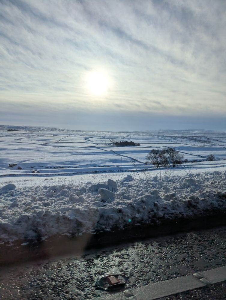 Fields covered in snow seen from a roadside edged with piled up snow and mud; sun shines dimly from behind fluffy white clouds 