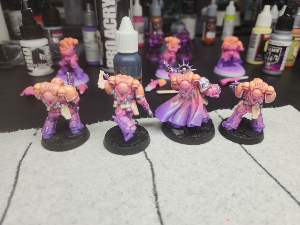 A group of 4 painted Warhammer 40k Space Marines. They are painted in a gradient of orange, pink, and purple with silver and white details.
They sit on a painting desk with other in progress minis and paints scattered about.