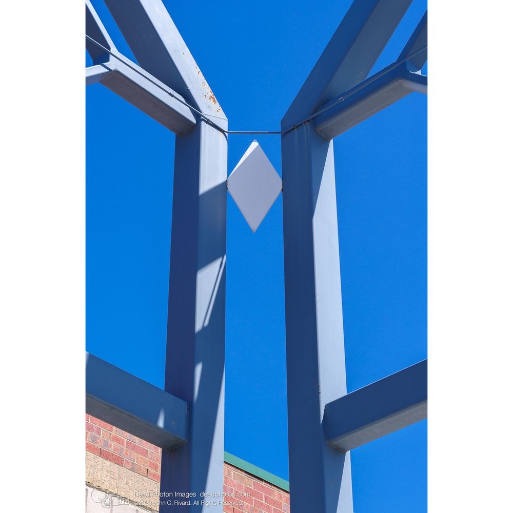 A decorative white diamond-shaped metal plate joins the corner where two vertical beams meet of a clock tower meet. A conduit pipe passes above it. At the lower left, the top of a red brick building wall is seen. Beyond a cloudless blue sky shows through the structure.