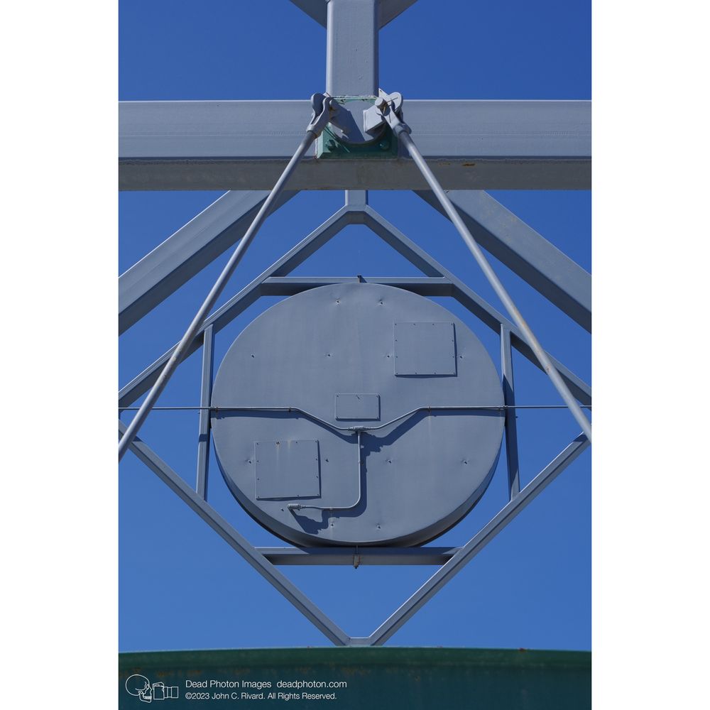 A grey-blue metal circle with screwed-on access plates (the back of a large clock) is surrounded by diamond-shaped steel beams and support shafts. Beneath is a green rounded girder.  Beyond a cloudless blue sky shows through the structure.