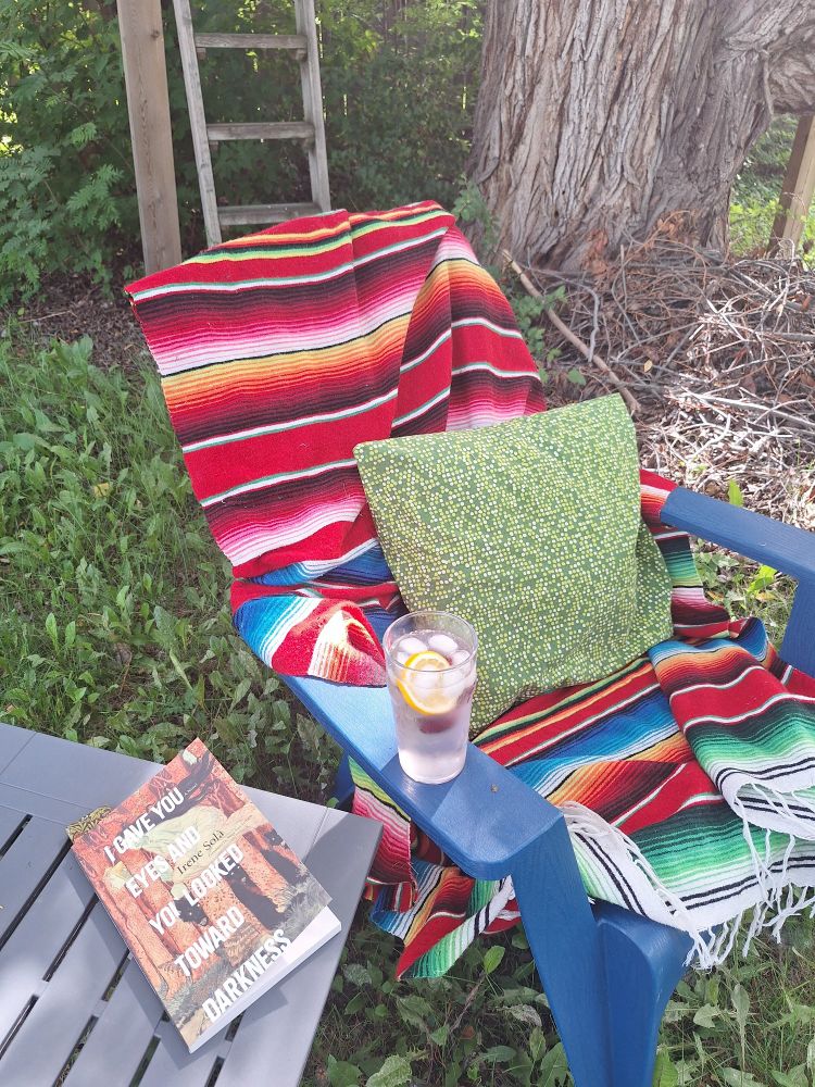 Garden scen with chair covered in colourful blanket and cushion, an iced drink on the arm. Beside it, a table carries a copy of 'I Gave You Eyes and You Looked Toward Darkness' by Irene Sola.