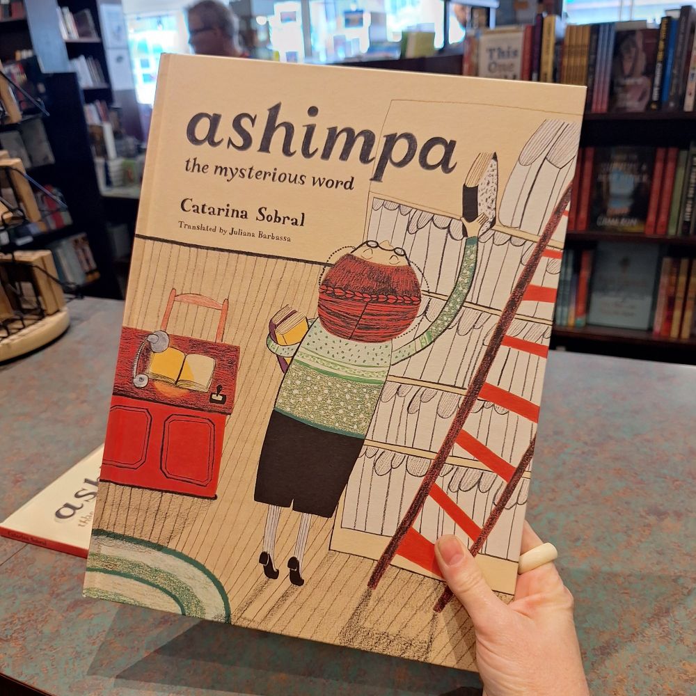 A hand holds up a children's picture book title 'ashimpa the mysterious word' by Catarina Sobral.