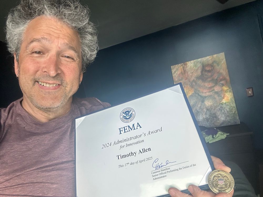 Gray-haired smiling man holding a framed award from FEMA and a challenge coin. 
