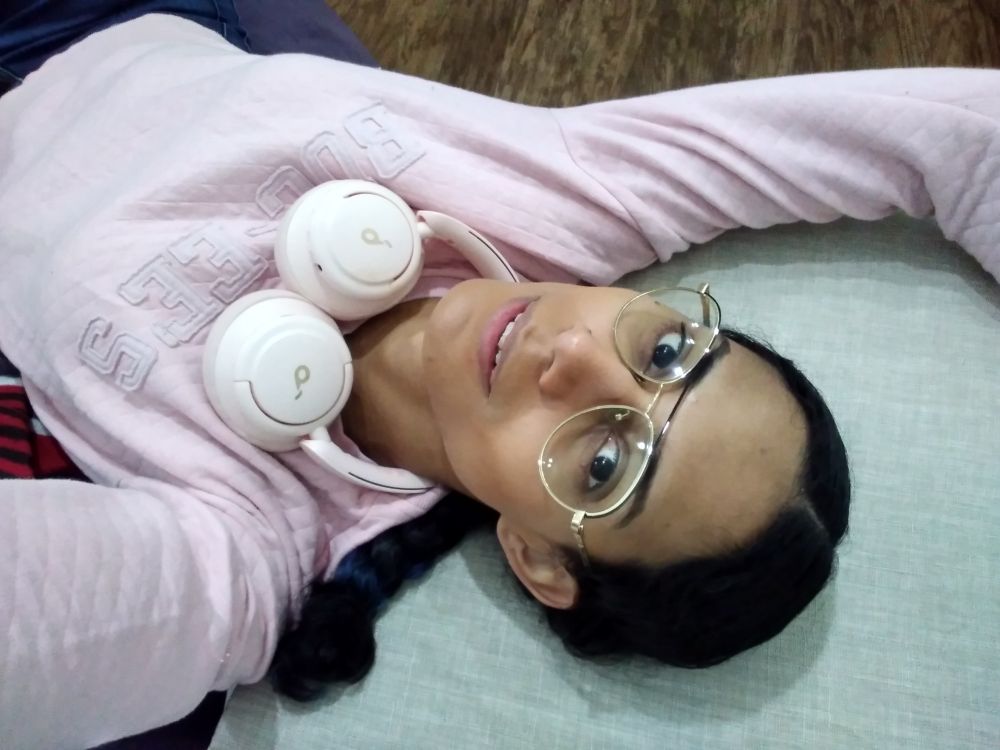 Sideways selfie of a latina sitting on a beanbag while resting her head on an off-white couch. She has center-parted black hair in a braid, golden aviators with clear lenses, pink headphones resting around her neck, and a matching pale pink Buc-ee's sweater.

One arm rests out of frame on the cushion above her head while the other stretches out to the camera. Her lips are parted in an inviting half-smile. There is a wooden floor pattern in the background.