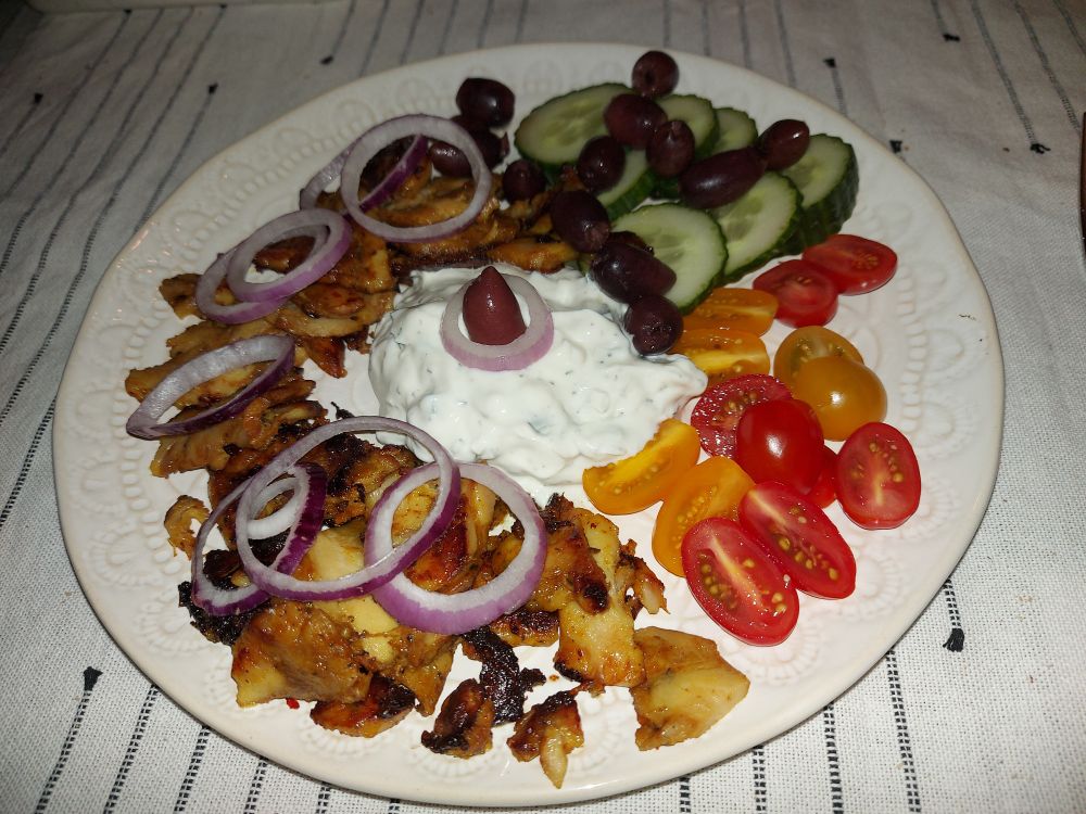 Heller Teller, darauf Hähnchengyros, Zwiebelringe, Tsatsiki, Tomaten, Gurkenscheiben und Oliven.