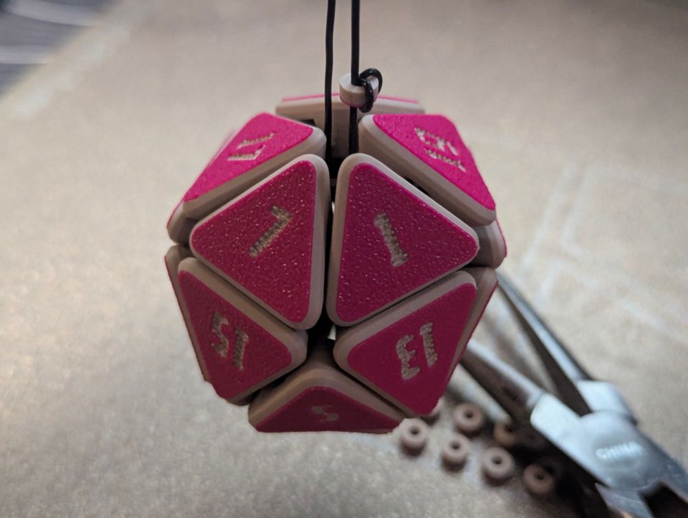 A 3d printed flexy D20 hanging from an elastic thread. Pliers and 3d printed beads in the background.