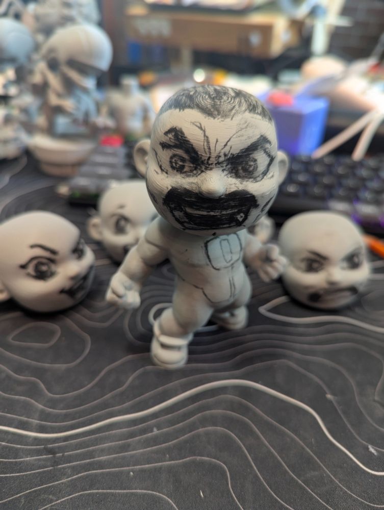 A raw, unpainted 3D-printed figure in a heroic stance with clenched fists, printed on a Bambu Labs X1Carbon FDM printer. A few other heads with different expressions on them are lying around. The gray plastic surface is covered in hand-drawn charcoal pencil sketches outlining Omni-Man’s rage filled face, and costume details, including a chest emblem, and costume. Light shading marks muscle definition on the torso and limbs. The minimalist design—with smooth layers and slight FDM texture—showcases the blank-canvas potential of the Li’l FAB•!T series by HarperCrafthall, meant for artists to plan, sketch, and paint their own custom characters.