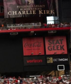 A crop of an image from the stadium during Charlie Kirk’s memorial showing a banner saying “Building a Legacy: Remembering Charlie Kirk” and then just below it are banners advertising Coca-Cola, Seat Geek, Verizon, and Bet MGM.
