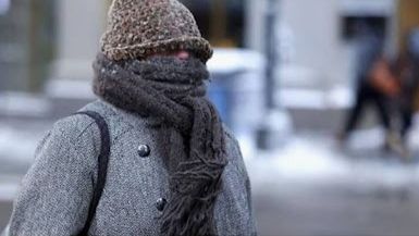 Foto de pessoa na rua completamente encasacada, de gorro e cachecol tudo em tons de cinza, só se vê o nariz de fora.