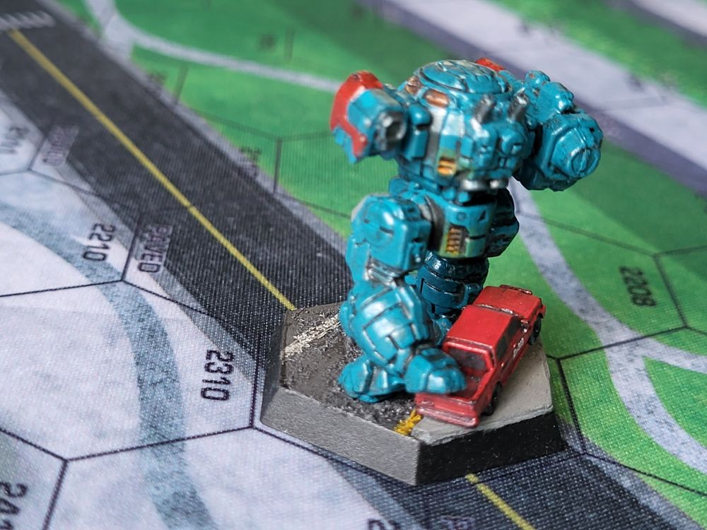 A teal blue Urbanmech steps on the tailgate of a red civilian truck. The truck is painted with D-319 in white letters on the passenger door. The end of the Urbanmech's massive autocannon is painted red and marred with carbon. (Painted miniatures)