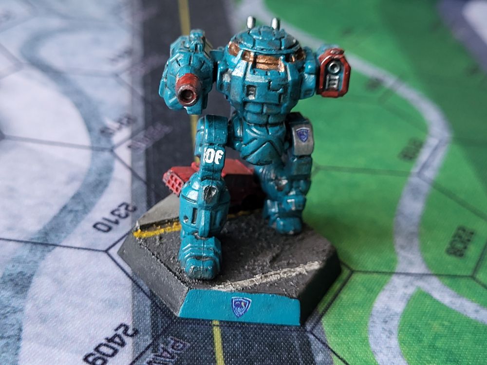A teal blue Urbanmech steps on the tailgate of a red civilian truck. The truck is painted with D-319 in white letters on the passenger door. The end of the Urbanmech's massive autocannon is painted red and marred with carbon. The left thigh bears the crest of the Free Rasalhague Republic. The right thigh bears the scratched up designator 06. (Painted miniatures)