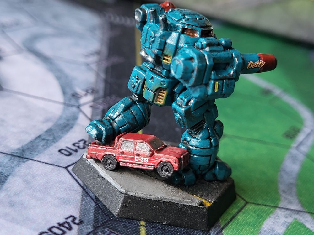 A teal blue Urbanmech steps on the tailgate of a red civilian truck. The truck is painted with D-319 in white letters on the passenger door. The Urbanmech's massive autocannon is decorated with the name Betty. The end of the autocannon is painted red and marred with carbon. (Painted miniatures)