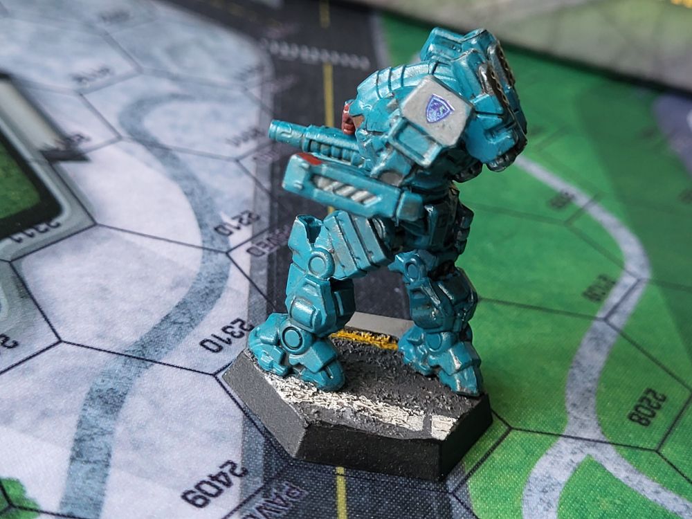 A teal blue Guillotine battlemech stands on a city street, ready for combat. It's hands are painted red in remembrance of the blood shed for Rasalhague independence. The emblem of the Free Rasalhague Republic is proudly displayed on its left shoulder. (Painted miniatures)