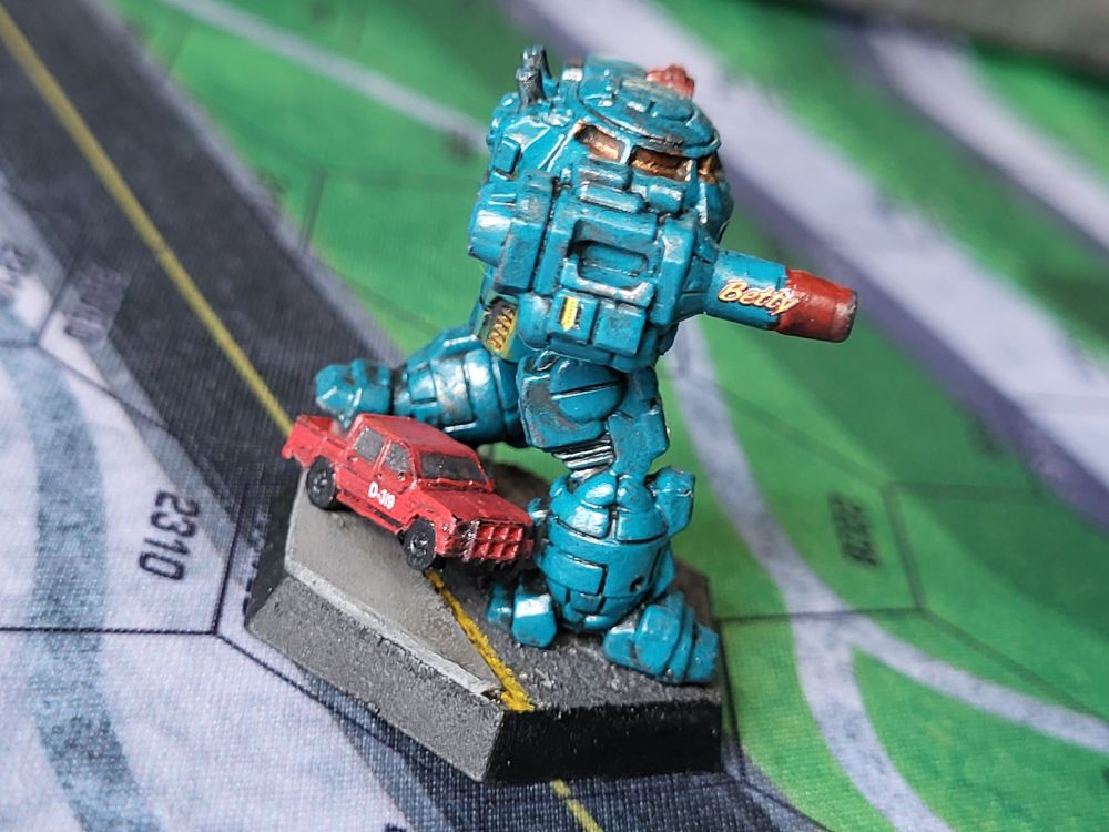 A teal blue Urbanmech steps on the tailgate of a red civilian truck. The truck is painted with D-319 in white letters on the passenger door. The Urbanmech's massive autocannon is decorated with the name Betty. The end of the autocannon is painted red and marred with carbon. (Painted miniatures)