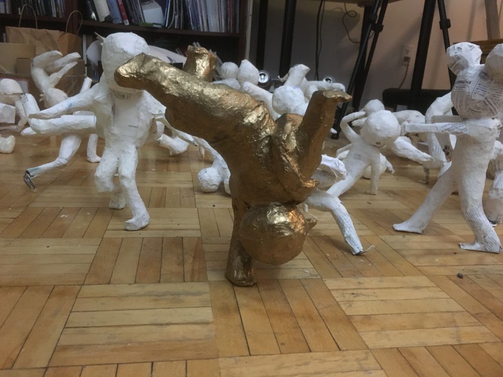 A golden break dancing sculpture balancing upside down on one hand, placed on a hardwood floor, with a collection of other break dancing sculpture characters (about 20) behind it.