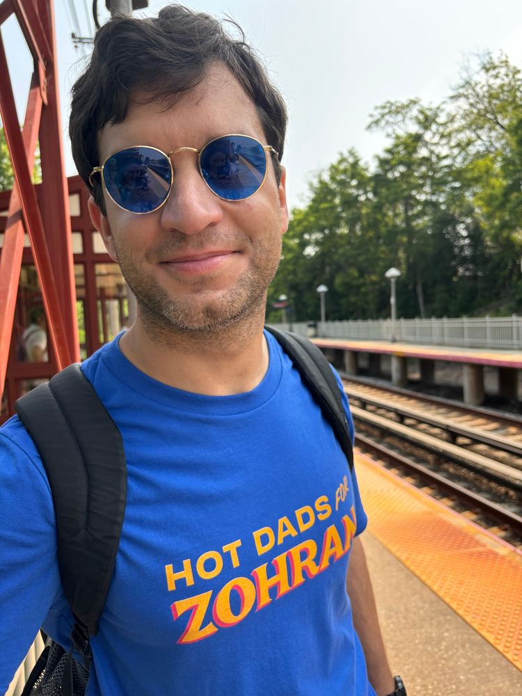 Adam Lee (@daylightatheism.bluesky.social) wearing a “hot dads for Zohran” shirt standing on an outdoor train platform 