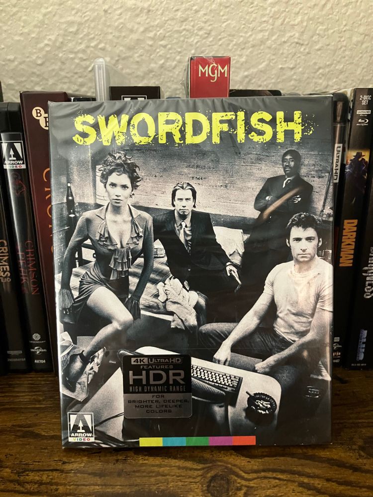 Front slipcover of Arrow Video’s 4K Ultra HD release of Swordfish. The artwork features a black-and-white, stylized photo of the main cast — Halle Berry, John Travolta, Don Cheadle, and Hugh Jackman — posed in a gritty, industrial setting. The title Swordfish is printed in bold, distressed yellow lettering at the top. A 4K HDR sticker is placed on the plastic wrap, and the Arrow Video logo is visible in the bottom corner. The slipcover appears to be sealed and in excellent condition, displayed on a shelf with other collector titles.