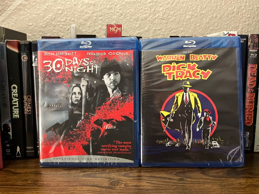 Two Blu-ray cases standing upright on a shelf. The left is 30 Days of Night, featuring a gritty, blood-spattered cover with Josh Hartnett and Melissa George in monochrome red and black tones. The right is Dick Tracy, showcasing bold comic-style artwork of Warren Beatty in a yellow trench coat and fedora, walking with a young boy against a dark background with a red circle and vintage cars. Both cases are still sealed in plastic wrap, surrounded by other Blu-ray titles on a wooden shelf.