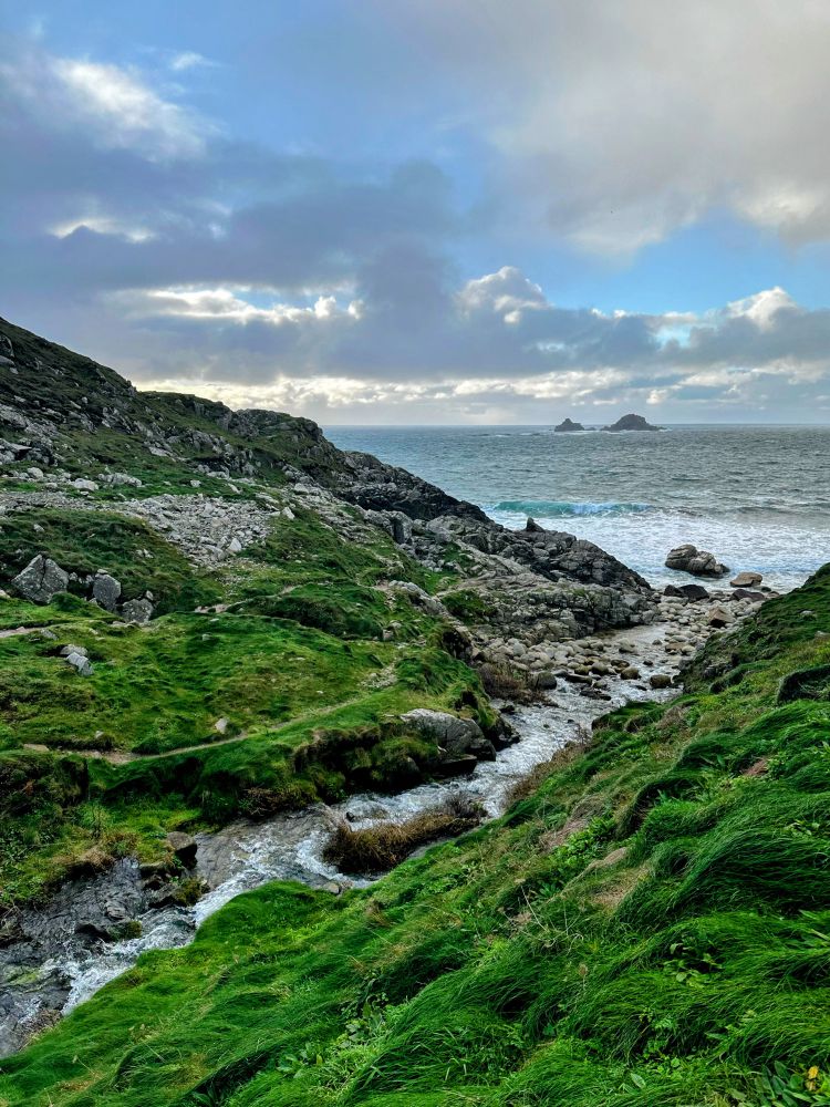 A fast-flowing steam enters the sea at a boulder-strewn cove in far west Cornwall. There are rocky islets on the horizon. The blue sky is heavy with big grey and white and clouds, and a weak light as it is the end of the day.