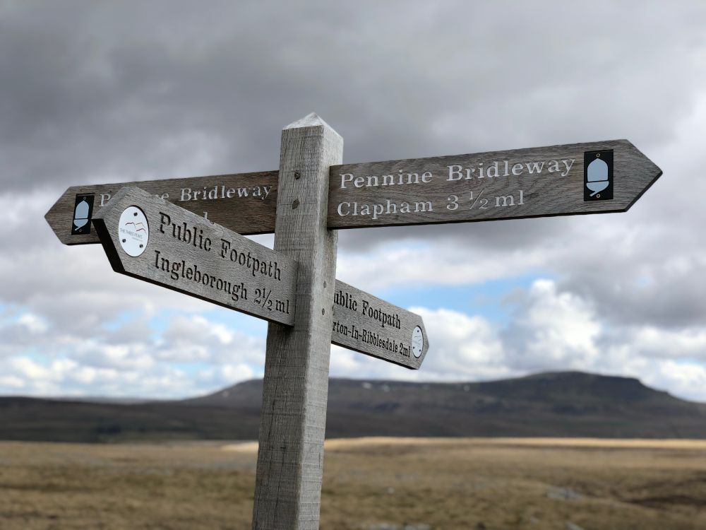 A four-way signpost. The Pennine Bridleway runs left-right. Ahead is the footpath to Horton-in-Ribblesdale and the outline silhouette of Pen-y-Ghent mountain. Behind camera would be Ingleborough, the sign says so.