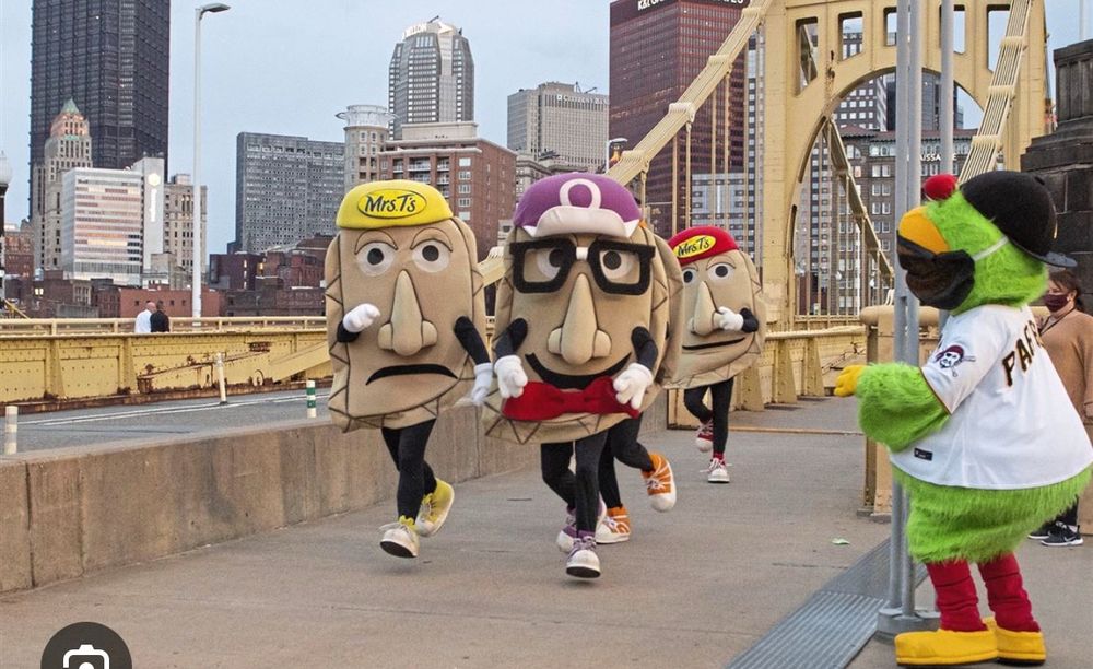 A picture of the Pittsburgh Pirates racing Pierogis. Four anthropomorphic pierogies (people in costumes) run over the gold-colored Clemente Bridge. On the left, a pierogi in yellow. In the center, a pierogi in purple with glasses. Behind, just showing feet, a pierogi in orange. Back right, a pierogi in red. The Pittsburgh Parrot cheers them on to the right