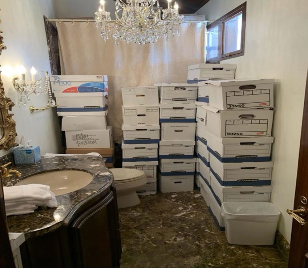 A photo taken by the Department of Justice in 2021. It is of cardboard file boxes piled up in a Mar-a-Lago bathroom. To the left is a sink with dark red and brown stone top and mirror with gold filigree. There is then an off-white toilet, chandelier and crystal-spangled wall light fixture. Stacked in front of a bathtub, continuing to the right wall are so many boxes