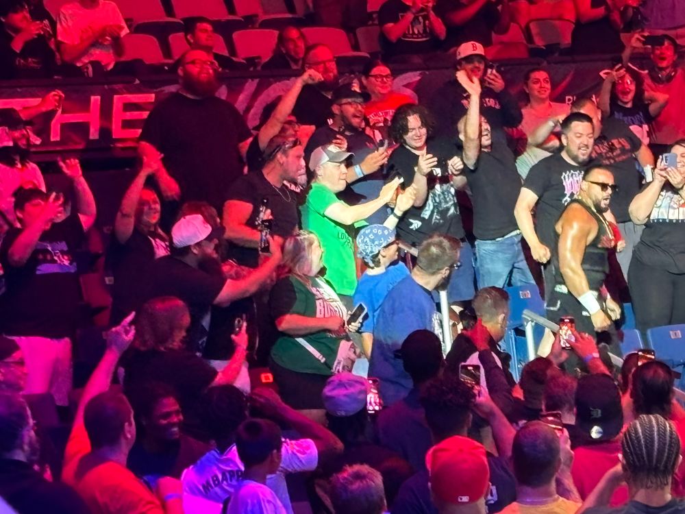 A picture of professional wrestler Mike Santana coming through the crowd. He is on the right side, in a spotlight, while the crowd cheers and takes pictures. Santana is wearing sunglasses, has black hair pulled back in a braided ponytail with a short black beard and wears a black tank and pants