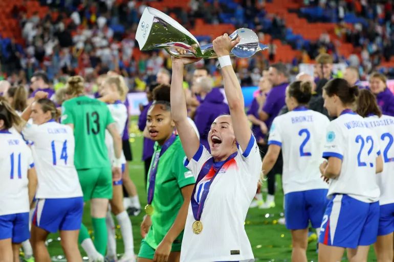 Chloe Kelly in an England jersey, mouth open in triumph, surrounded by teammates she hoists the UEFA Women’s Euro 2025 trophy under stadium lights.